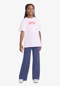 Λευκό μπλουζάκι με ροζ λογότυπο "Converse", συνδυασμένο με παντελόνι φαρδιάς γραμμής σε σκούρο μπλε και ανοιχτό ροζ παπούτσια. Πλεξούδες μαλλιών πέφτουν στους ώμους.