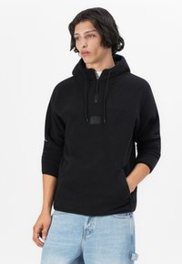 Pullover in pile nero con cappuccio, zip mezza lunghezza, tasche a marsupio frontali e polsini elasticizzati. Presenta un patch con logo sul petto. Vestibilità casual.