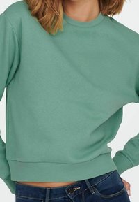 Žena v jednobarevném svetru v mořské zelené barvě s krátkým rukávem a tmavě modrých džínách, s rukama na bocích, na bílém pozadí.