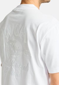 Weißes Baumwoll-T-Shirt mit geprägten Grafiken auf der Rückseite, einschließlich Text und einem runden Logo, mit rundem Ausschnitt und kurzen Ärmeln.