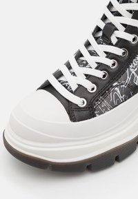 Sneaker alta con un motivo grafico in bianco e nero, pelle nera liscia, lacci bianchi e una suola robusta in gomma bianca e nera.