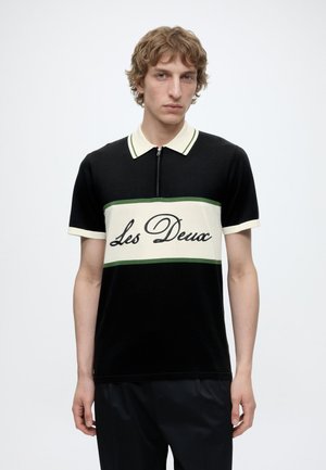 Jeune homme aux cheveux clairs et bouclés, portant un polo noir à manches courtes avec des rayures horizontales crème et vertes et le texte « Les Deux » sur la poitrine.