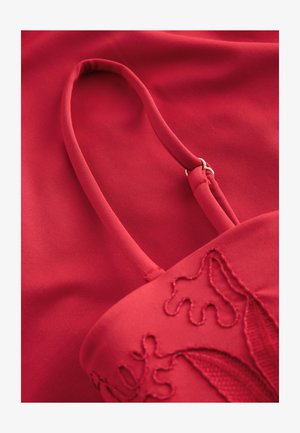 Rote Stofftasche mit einem strukturierten, bestickten Design und einem flachen Riemen, der durch einen Metallring gesichert ist, vor einem glatten roten Hintergrund.