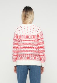 Mulher vestindo um suéter de malha com padrão branco e vermelho e jeans azuis, de costas para a câmara, contra um fundo claro e liso.