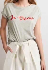 Light gray short-sleeve T-shirt featuring red embroidered text "Je t'aime." Worn with white high-waisted pants tied at the waist.