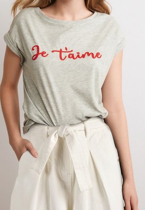 Un T-shirt gris clair à manches courtes avec un texte brodé rouge "Je t'aime." Porté avec un pantalon blanc taille haute attaché à la taille.