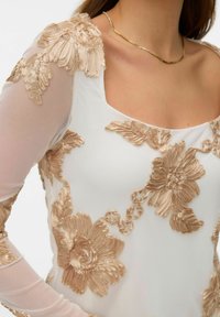 Bílý průhledný top s dlouhými rukávy zdobený velkou,texturovanou zlatou květinovou výšivkou a čtvercovým výstřihem, nošený se zlatým řetízkovým náhrdelníkem.