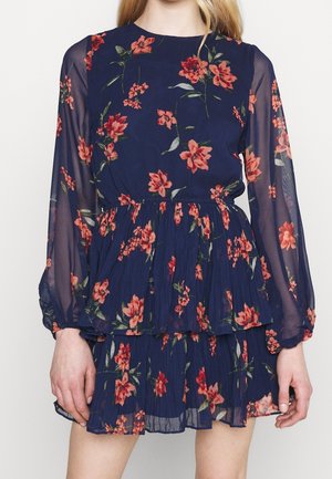 Femme portant une robe bleu marine à manches longues avec des manches transparentes et un imprimé floral rouge, avec une jupe à étages et une taille élastique.