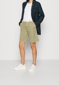 Nainen, joka pukeutuu vihreään ruudulliseen bleiseriin, valkoiseen paitaan, beigeihin shortseihin, joissa on kudottu vyö, ja valkoisiin lenkkareihin, seisoo kädet taskuissa.