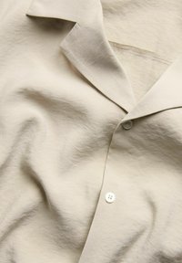 Beige Knopfhemd aus einem weichen, strukturierten Stoff. Verfügt über einen Reverskragen, einen einzelnen weißen Knopf und einen dezenten Glanz.