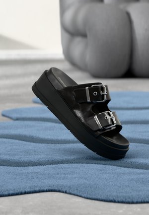 Sort slidesandal med to sølvspænder, platårsål, placeret på tekstureret blåt tæppe med slørede beige møbler i baggrunden.