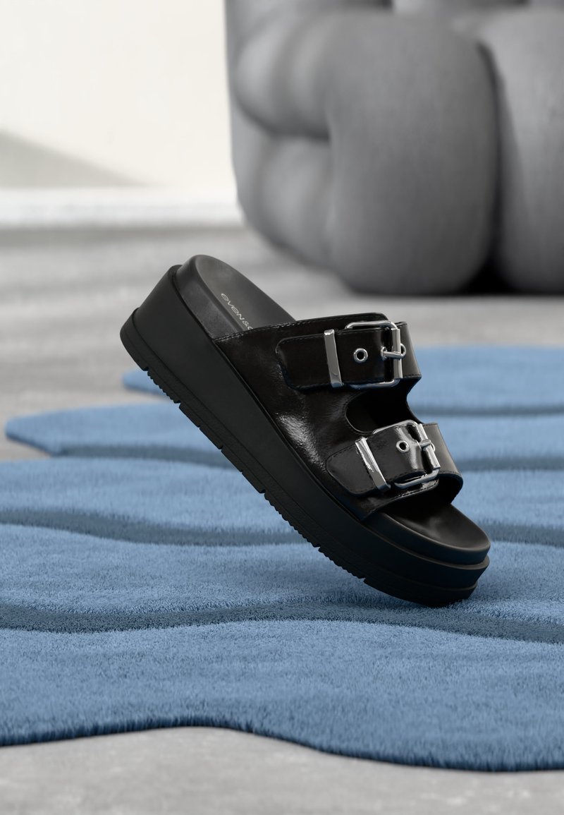 Black slide sandal with two silver buckles, platform sole, placed on textured blue rug with blurred beige furniture in background.