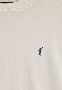 Cremefarbener Pullover mit einem gerippten Rundhalsausschnitt und einer kleinen schwarzen bestickten Figur, die auf der linken Seite eine Tasche trägt. Weicher, strukturierten Stoff.