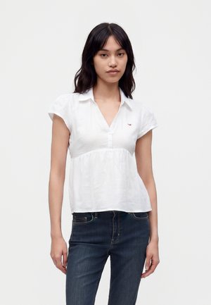 Junge Frau mit dunklen Haaren, die eine kurzärmelige weiße Bluse und dunkelblaue Jeans trägt und vor einem einfarbigen hellen Hintergrund steht.