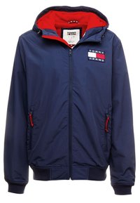 Navy-Jacke mit roter Kapuze, vorn mit Reißverschluss, zwei seitlichen Taschen mit roten Akzenten und einem Logo-Patch auf der Brust. Leichtes Material.