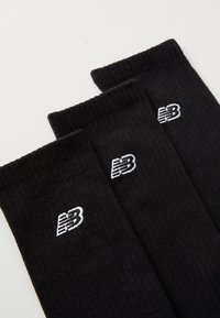 Calcetines de algodón negros con textura acanalada y logo de New Balance bordado en blanco. Tres calcetines están apilados.