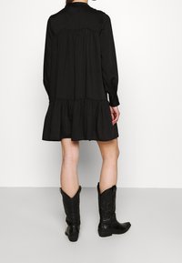 Un vestido negro de manga larga presenta un diseño arrugado con un dobladillo en niveles. Se lleva con botas de cowboy negras, mostrando una textura suave y un ajuste relajado.