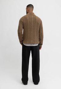 Brauner gestrickter Pullover mit Kabelmuster, hohem Kragen, gerippten Bündchen und Saum. Kombiniert mit lockeren schwarzen Hosen und Schuhen.