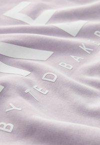 Světle fialová látka s bílým textem „BY TED BAKER“. Textura vypadá jemně a hladce s jemným leskem.