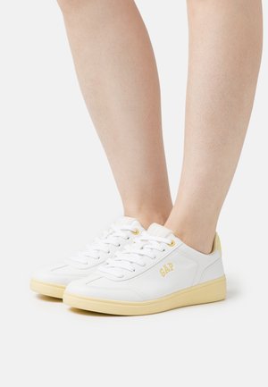 Sneakers bianchi con accenti gialli, caratterizzati da un design in pelle texturizzata, punta rotonda, chiusura con lacci e logo "GAP" sul lato.