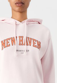 Felpa leggera di colore rosa con tasca a marsupio, cordini e testo arancione "NEW HAVEN SPORTS CLUB." Tessuto morbido, vestibilità rilassata e polsini a costine.