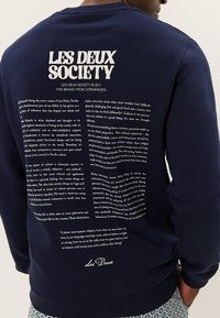Sudadera de color azul marino con texto blanco en la parte posterior, que incluye un nombre de marca y un bloque de texto, con dobladillo redondeado y puños acanalados.