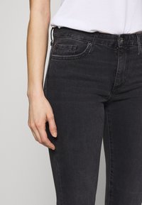 Černé džíny ze denimu s přiléhavým střihem. Mezi vlastnostmi jsou přední kapsy, poutka na opasek a zip na přední straně. Materiál vypadá hladce s jemnou strukturou.