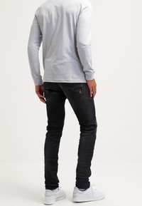 Man wearing light grey long-sleeve shirt, black jeans, and white sneakers standing facing away against plain white background.