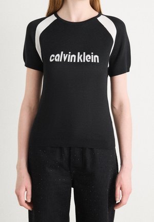 Person iført en sort kortærmet top med hvide skulderpaneler og teksten "calvin klein" foran, kombineret med sorte bukser.