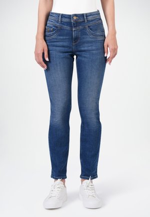 Slim-Fit blaue Jeans, getragen von einer stehenden Person mit weißen Sneakern vor einem einfarbigen Hintergrund.