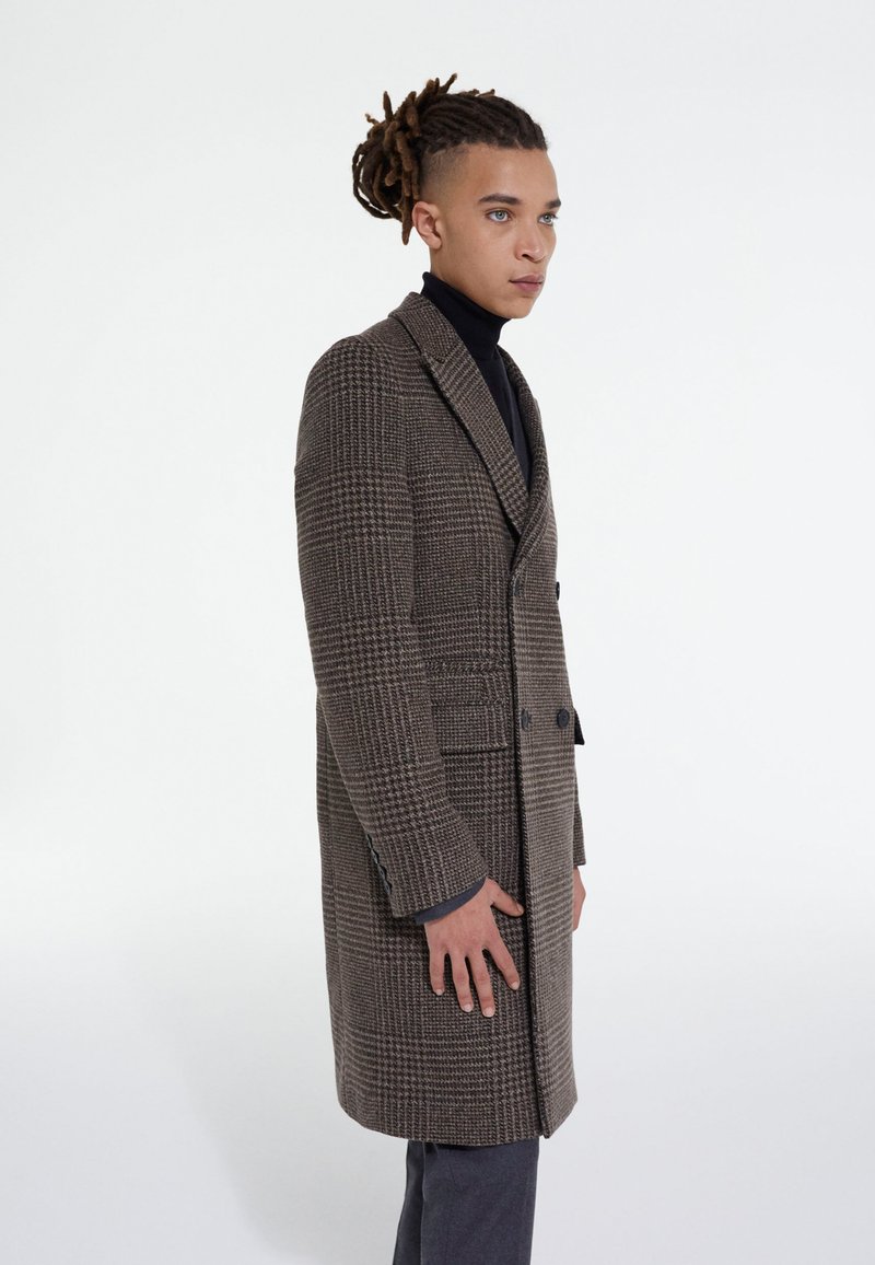 Cappotto lungo a quadri marroni con ampie rever, bottoni doppi e tasche laterali. Indossato sopra un dolcevita nero. Tessuto liscio.