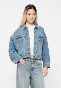 Veste en denim coupée de couleur bleu clair avec une coupe décontractée, fermeture à boutons, deux poches avant et manches larges. Associée à un jean taille haute.
