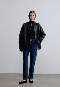 Μαύρο μπουφάν bomber από ψεύτικο δέρμα με ψηλό γιακά, φορεμένο πάνω από μαύρη μπλούζα με μακριά μανίκια, συνδυασμένο με μπλε τζιν και μαύρα παπούτσια χωρίς κορδόνια.