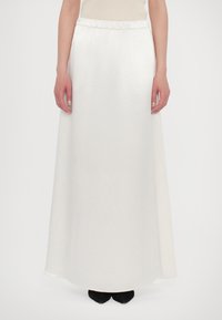 White maxi skirt with a gathered waistband, smooth texture, and a flowing design, reaching the ankles. Paired with black footwear.