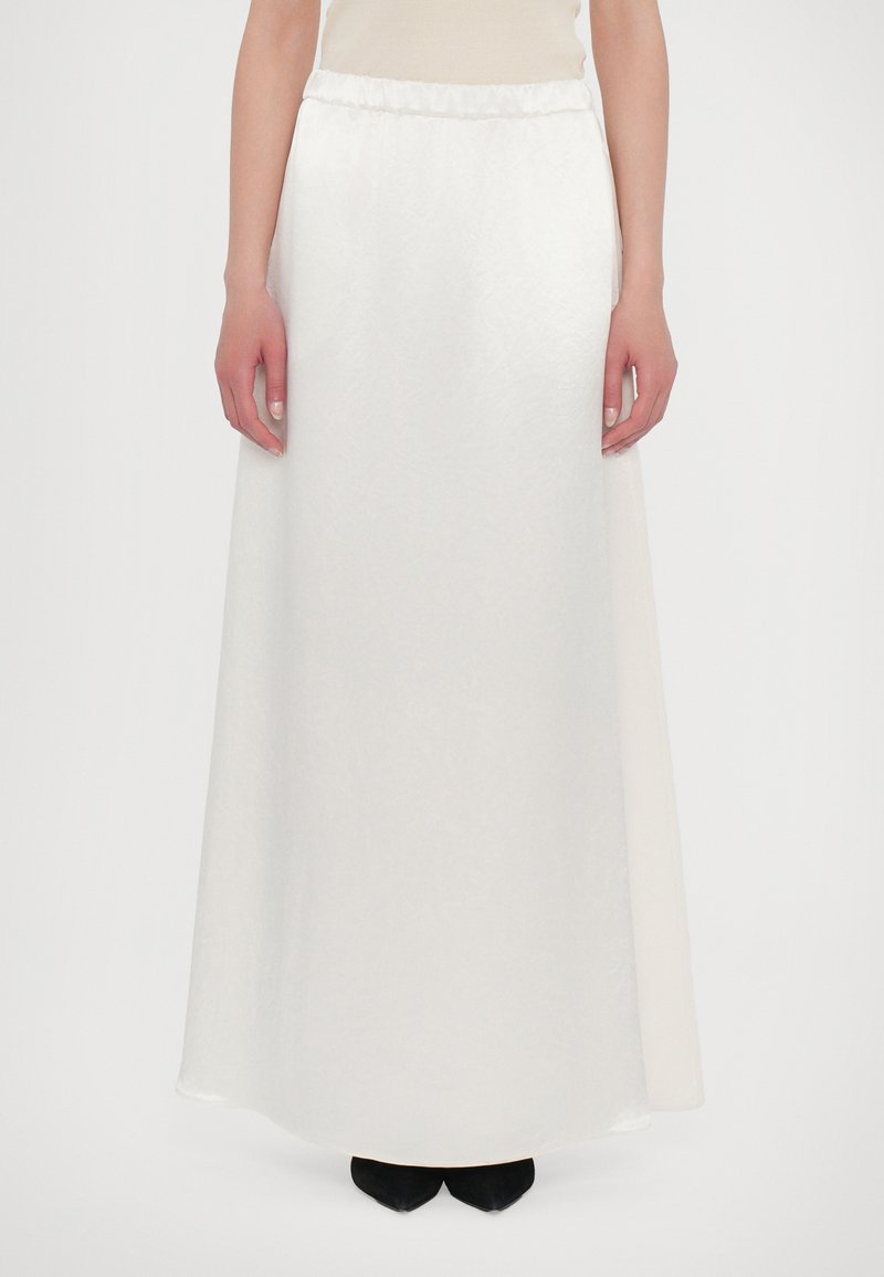 White maxi skirt with a gathered waistband, smooth texture, and a flowing design, reaching the ankles. Paired with black footwear.