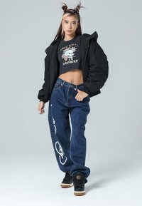 Chaqueta acolchada negra, camiseta gráfica negra ajustada con un diseño de calavera, jeans azules sueltos con letras blancas y zapatillas negras con suelas marrones.