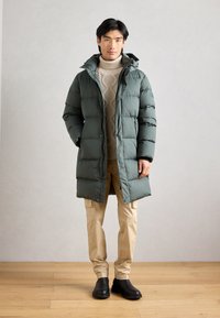 Vihreä puhvitakki hupullinen, tikattu pinta, edessä napituskohdat, päällä kerma-värinen palmikkoneule ja vaaleat housut. Mustat kengät.