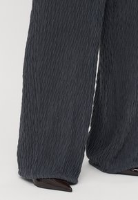 Pantalons à jambes larges en tissu texturé gris foncé, présentant un design froissé et un ourlet se terminant juste au-dessus de talons aiguilles noirs.