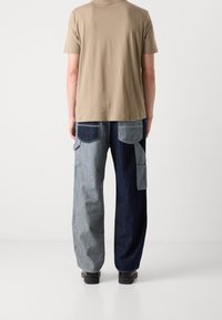 Mand i tofarvede denim cargo bukser med mørkeblå og lyseblå paneler, beige kortærmet skjorte og sorte sko, stående med ryggen til.