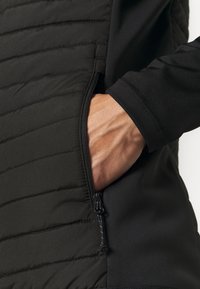 Schwarze Jacke mit einem seitlichen Reißverschlussfach und gestepptem Design. Aus strapazierfähigem Stoff mit glatter, matter Oberfläche und enganliegenden Bündchen.