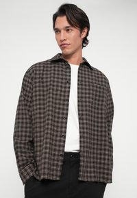 Giovane uomo che indossa una camicia a quadri marrone e beige sopra una maglietta bianca, con pantaloni neri, che guarda leggermente di lato.