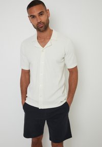 Strik kortærmet skjorte i off-white med et struktureret mønster, der har V-udskæring og frontknaplukninger, parret med marineblå shorts.