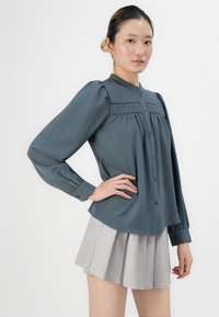 Kvinne med hår i en knute iført en grå bluse med langt ermet og knapper, samt en lys grå plissert skjørt mot en hvit bakgrunn.