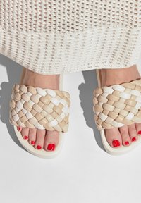 Woven sandals with a beige and white design, featuring a wide strap and smooth soles, paired with red polished toenails.