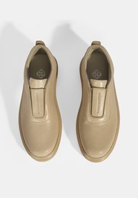 Beige instapschoenen van geperforeerd leer met een gladde neus en een ronde achterkant, voorzien van een opvallend centraal paneel en een gestructureerd oppervlak.