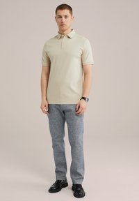 Camisa polo beige de manga corta y cuello, combinada con pantalones texturizados gris claro y zapatos negros, con tres botones en el cuello.