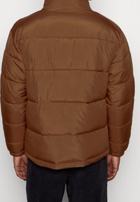 Brązowa kurtka puchowa z gładką, błyszczącą powierzchnią, pikowanym wzorem, wysokim kołnierzem i elastycznymi mankietami. Kurtka ma luźny krój.