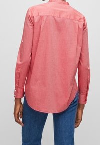 Person iført en langærmet koralrød skjorte med knapper og blå jeans, vist fra ryggen mod en ensfarvet lys baggrund.