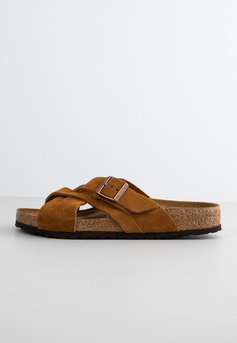 Sandale à glissière en daim marron avec des straps croisées et une boucle métallique. Semelle intérieure en liège texturé et semelle extérieure en caoutchouc noir.