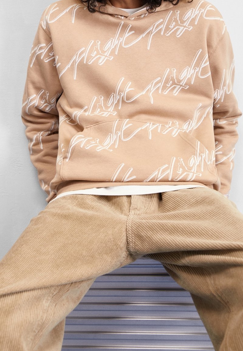 Beige mikina s bílými křivkovými grafikami "flight", má přední kapsu a kapuci se stahovacími šňůrkami. Nošena s béžovými kordové kalhoty.